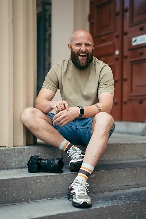 Een fotograaf zwolle zit op een trap te lachen met zijn camera voor zich.
